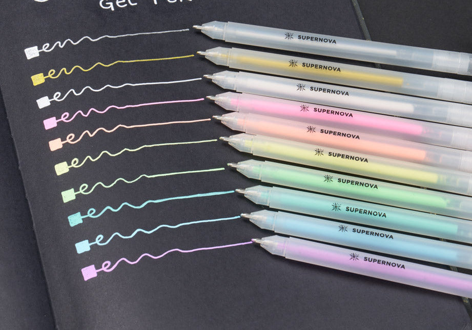 A black notebook shows nine gel pens labeled "SUPERNOVA," each drawing a colorful line. The lines display pastel colors on black paper, highlighting the pens' vibrant hues.